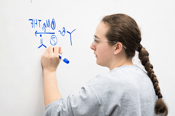 A student writes a chemical formula on a whiteboard with her blue marker.